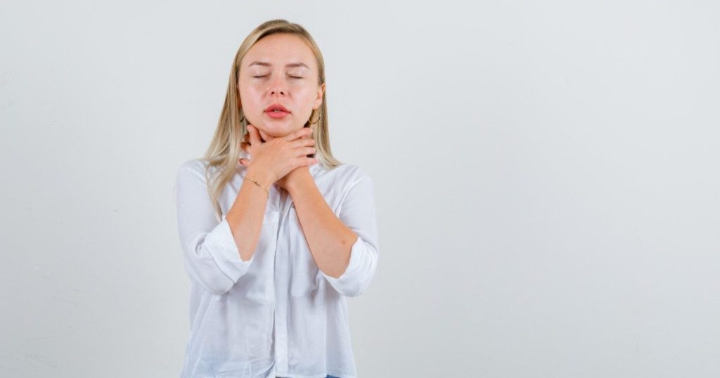 A woman holding her throat with both hands, eyes closed and wearing a slightly pained expression due to tonsil stones